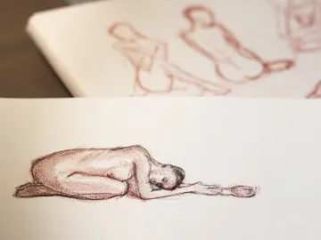 Nackte Figur liegt liegend auf Papier, umgeben von Skizzen.